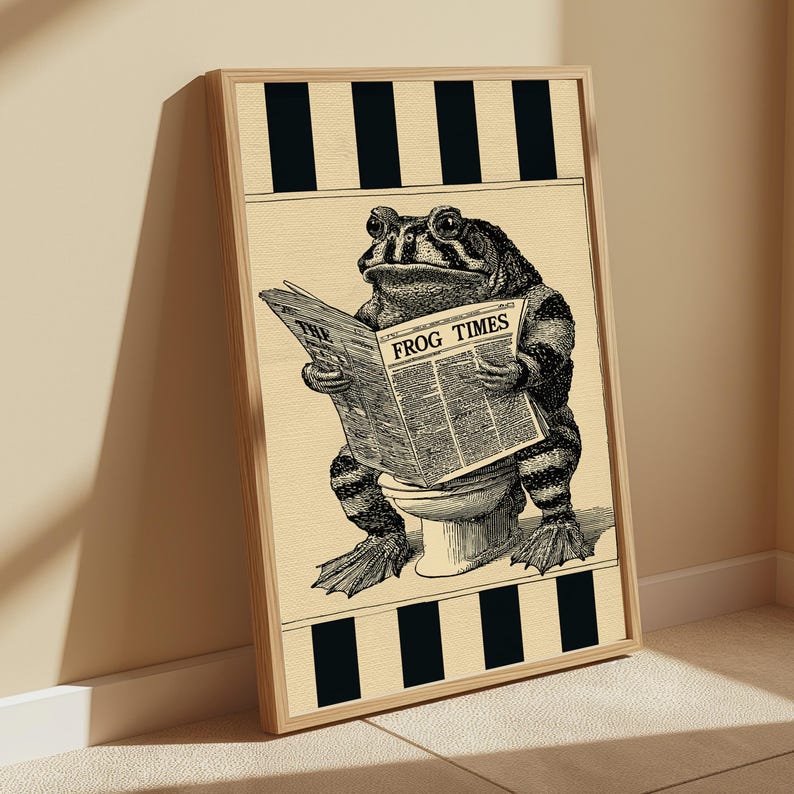 May include: A framed wall art print featuring a black and white illustration of a toad sitting on a toilet while reading a newspaper titled THE FROG TIMES. The background is a light cream colour with black vertical stripes at the top and bottom edges.