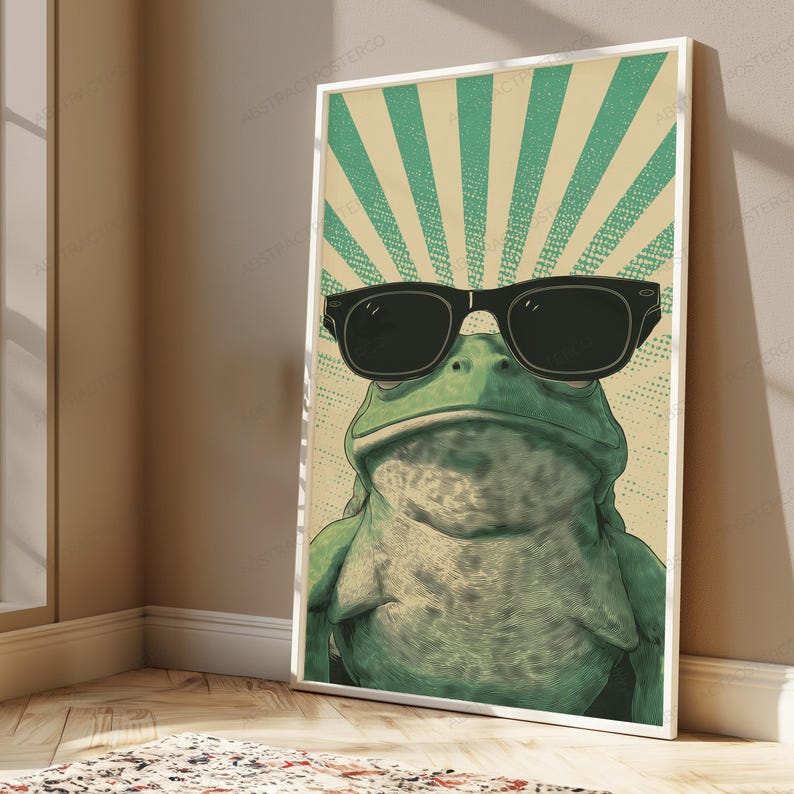 May include: A framed wall art print featuring a green frog wearing black sunglasses. The background consists of a light beige and teal green sunburst pattern with a dotted texture. The frog is centered, looking forward with a neutral expression.
