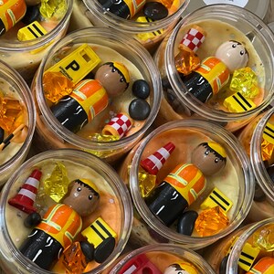 May include: Multiple clear jars filled with yellow and orange play dough, each containing a small wooden figurine wearing a yellow and orange construction worker outfit, along with other small toys like traffic cones, bricks, and gems. The jars are labeled "The Busy Play Box".
