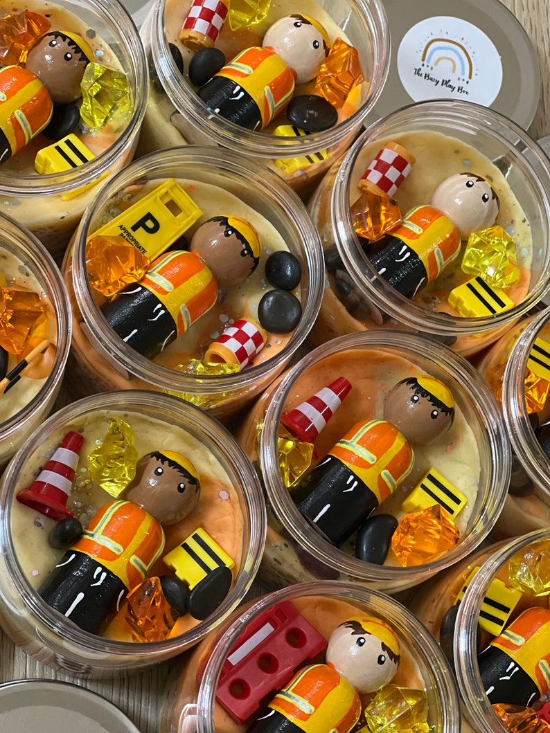 May include: Multiple clear jars filled with yellow and orange play dough, each containing a small wooden figurine wearing a yellow and orange construction worker outfit, along with other small toys like traffic cones, bricks, and gems. The jars are labeled "The Busy Play Box".
