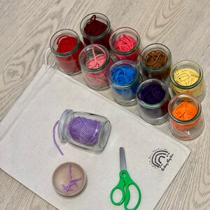 May include: A set of colorful yarn in glass jars, a small wooden bowl with purple yarn, a pair of green scissors, and a white canvas bag with the text "The Play Room" printed on it.