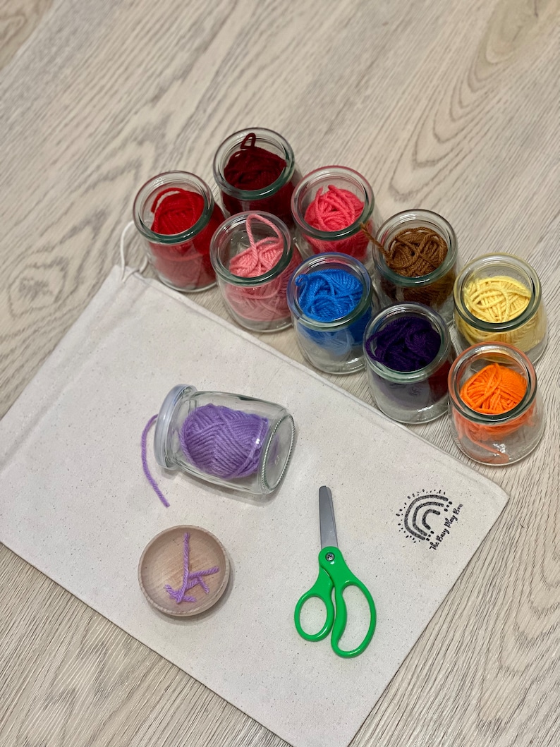 May include: A set of colorful yarn in glass jars, a small wooden bowl with purple yarn, a pair of green scissors, and a white canvas bag with the text "The Play Room" printed on it.