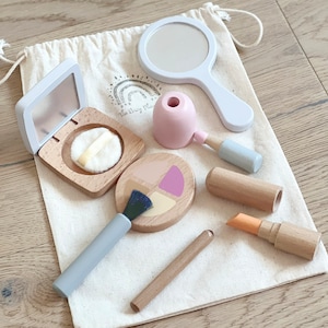 May include: A wooden toy makeup set in a white drawstring bag. The set includes a mirror, a compact with a powder puff, a blush palette, a lipstick, a lip gloss, and two brushes.