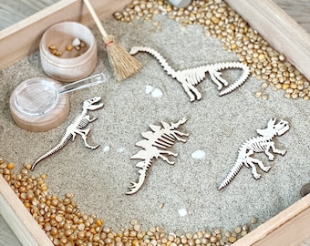 May include: A wooden tray filled with sand and small golden beads. There are four wooden dinosaur skeletons, a magnifying glass, a small wooden bowl, a broom, and two white eggs.
