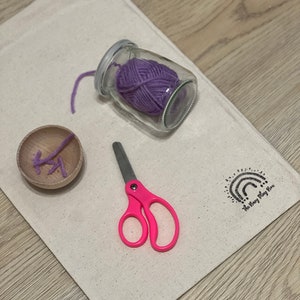 May include: A white canvas bag with a purple yarn ball in a glass jar, pink scissors, and a wooden spool with yarn. The bag has the text "The Story Play Box" printed on it.