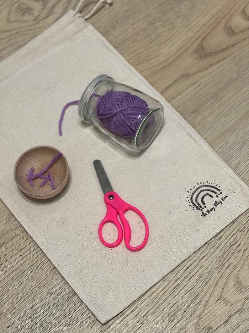 May include: A white canvas bag with a purple yarn ball in a glass jar, pink scissors, and a wooden spool with yarn. The bag has the text "The Story Play Box" printed on it.