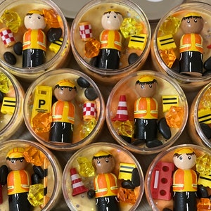 May include: Eight small glass jars filled with orange slime and various miniature construction-themed toys, including miniature construction workers, traffic cones, and road signs. The jars are arranged in a circle on a wooden surface.