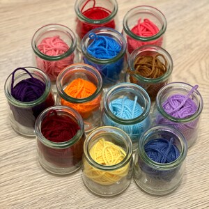 May include: A collection of 12 glass jars filled with colorful yarn balls. The jars are arranged in a circle on a wooden surface. The yarn colors include red, orange, yellow, green, blue, purple, pink, brown, and white.