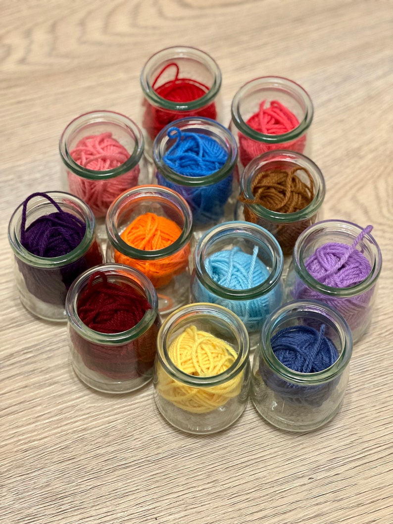 May include: A collection of 12 glass jars filled with colorful yarn balls. The jars are arranged in a circle on a wooden surface. The yarn colors include red, orange, yellow, green, blue, purple, pink, brown, and white.