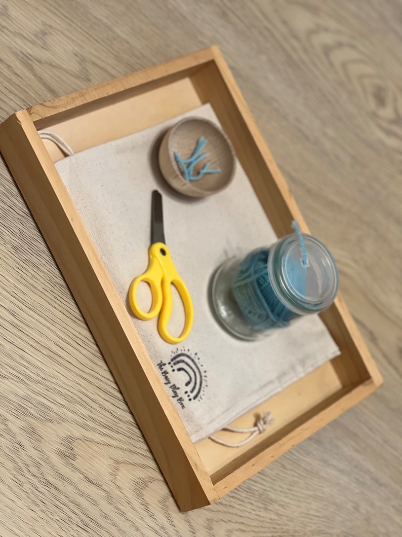 May include: A wooden tray with a white fabric bag, a small bowl of blue plastic pieces, a pair of yellow scissors, and a glass jar with blue string. The bag has the text "The Busy Play Time" printed on it.