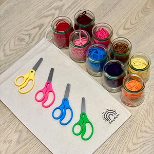 May include: A set of colorful yarn in glass jars, with four pairs of colorful scissors on a white canvas bag. The bag has a rainbow logo with the text "The Busy Play Box".