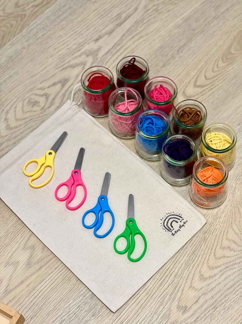 May include: A set of colorful yarn in glass jars, with four pairs of colorful scissors on a white canvas bag. The bag has a rainbow logo with the text "The Busy Play Box".