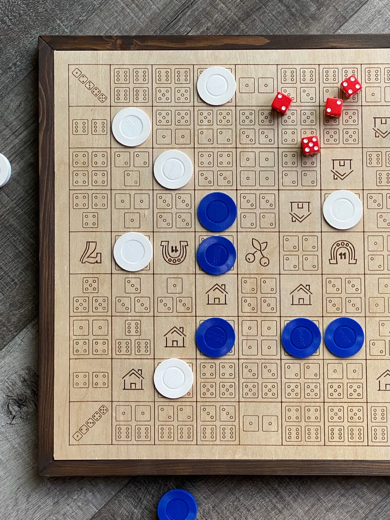 May include: A wooden board game with a dark brown frame. The game board has a grid pattern with various symbols and dice patterns. White and blue game pieces are scattered across the board, along with red dice.