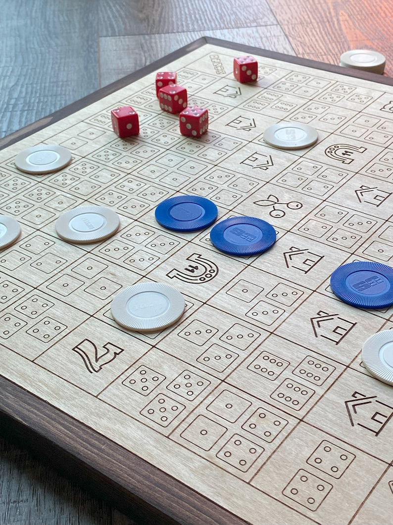 May include: A wooden board game with a dark brown frame. The light-colored game board features etched squares with dice patterns and symbols. Red dice and blue and white game chips are scattered across the board. A fun game for family and friends.