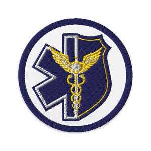 Puede incluir: Un parche bordado blanco y azul con un símbolo médico, una estrella y alas. El parche está rodeado por un círculo azul.