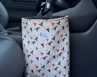 Puede incluir: Una bolsa de basura colgante para coche de tela blanca con un patrón repetitivo de sombreros de bruja, estrellas y lunas crecientes en naranja, negro y gris. Una pequeña etiqueta rectangular blanca con el texto negro Rafi Made está centrada.