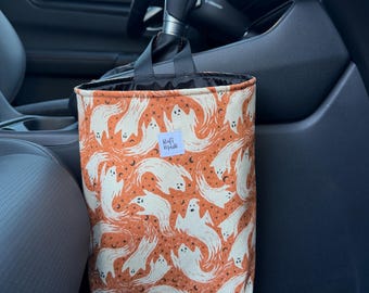 Puede incluir: Una bolsa de basura para coche de tela rectangular con fondo naranja y un patrón repetitivo de fantasmas blancos, pequeñas estrellas negras y lunas crecientes. Una pequeña etiqueta blanca con el texto Rafi Made está cosida al frente.