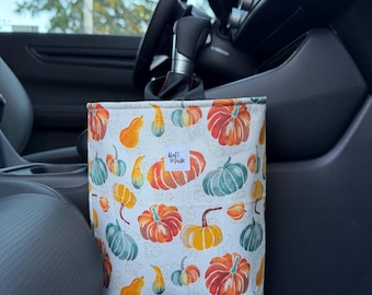 Puede incluir: Una bolsa de basura para coche de tela rectangular con un patrón repetitivo de calabazas naranjas, amarillas y verde azulado sobre un fondo blanco. Una pequeña etiqueta rectangular blanca con el texto negro "Rafi Made" está centrada cerca del borde.