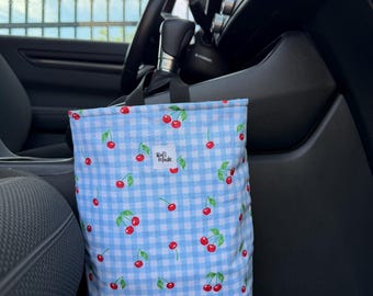 May include: A rectangular fabric car trash bag featuring a light blue and white gingham pattern with a repeating cherry print. A small white rectangular label with the text "Rafi Made" is sewn onto the front. The bag has a black strap.