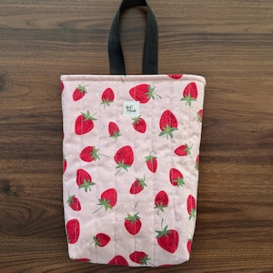 May include: A rectangular quilted fabric pouch featuring a light pink background with a repeating pattern of red strawberries and small white dots. The pouch has a black fabric loop handle at the top and a small white label reading Craft Made.