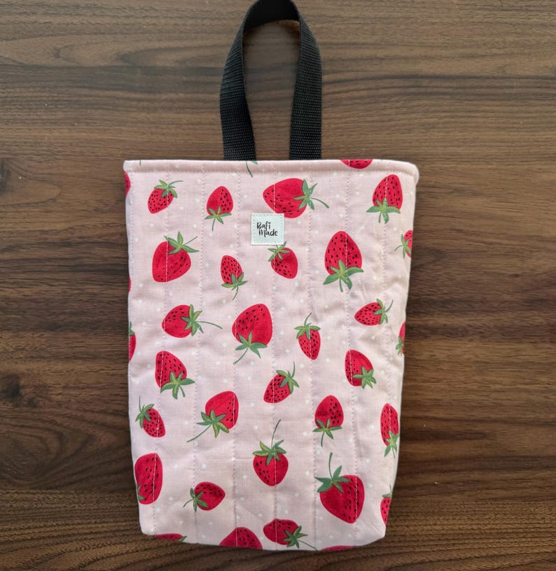 May include: A rectangular quilted fabric pouch featuring a light pink background with a repeating pattern of red strawberries and small white dots. The pouch has a black fabric loop handle at the top and a small white label reading Craft Made.