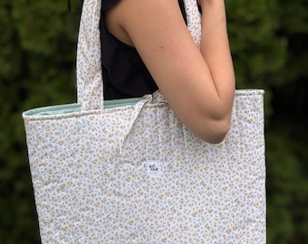 Puede incluir: Un bolso tote acolchado con un estampado floral a pequeña escala en amarillo y azul sobre un fondo blanco. El bolso tiene dos correas para el hombro y un forro interior de color verde claro. Incluye una etiqueta blanca con el texto Rafi Made.