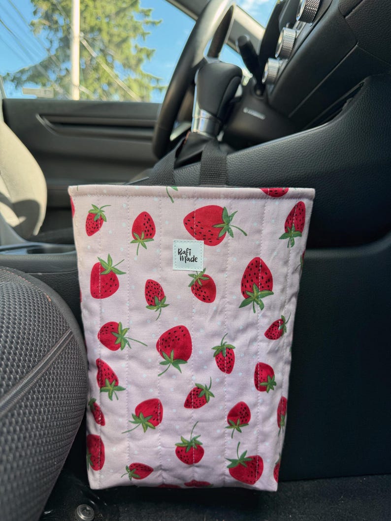 May include: A quilted fabric car trash bag featuring a light pink background with a repeating pattern of red strawberries and small white dots. The bag has black nylon straps and a small white rectangular label that reads Rafi Made in black text.