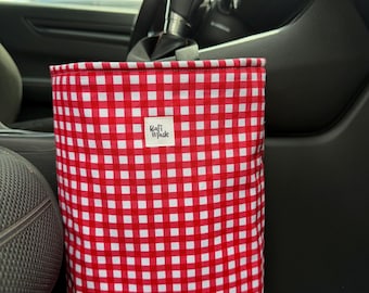 May include: A rectangular fabric car trash bag featuring a red and white gingham checkered pattern. A small white rectangular label with the black text Bafi Made is centered on the front of the bag. The bag is suspended from the vehicle gear shift.