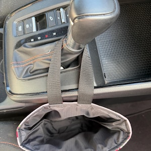 May include: A black fabric car trash bag with two black nylon straps hanging from a vehicle gear shift lever. The interior of the bag is lined with black material, and the exterior features a colorful floral pattern with green and pink tones.