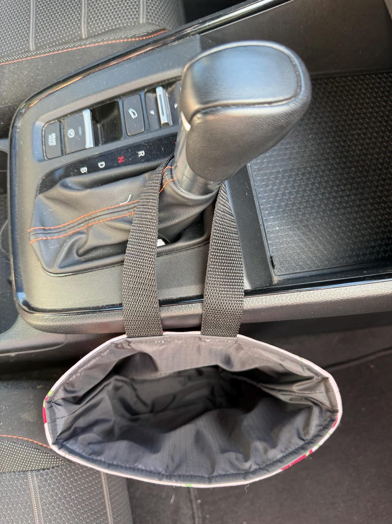 May include: A black fabric car trash bag with two black nylon straps hanging from a vehicle gear shift lever. The interior of the bag is lined with black material, and the exterior features a colorful floral pattern with green and pink tones.