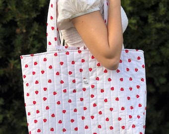 Puede incluir: Un bolso tote acolchado de color blanco con un patrón repetitivo de pequeñas fresas rojas. El bolso tiene líneas de costura verticales y una pequeña etiqueta de tela rectangular en el centro con el texto "Rafi Made" impreso en negro.