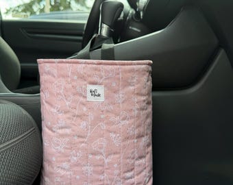 Puede incluir: Una bolsa de basura para coche rectangular y acolchada en tela rosa empolvado con un patrón floral de líneas blancas. La bolsa tiene un diseño de costuras verticales y una pequeña etiqueta de tela blanca con el texto Rafi Made.