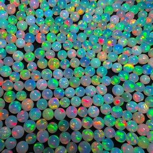 May include: A close-up of numerous iridescent opal gemstones. The opals exhibit a spectrum of colours, including green, blue, red, and yellow, with a milky white base. The gemstones are round and polished.