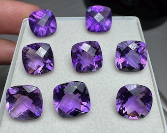 Könnte beinhalten: Sieben facettierte, quadratisch geschliffene Amethyst-Edelsteine in verschiedenen Lilatönen werden in einer weißen Schale präsentiert. Die Edelsteine haben einen Kissenschliff mit mehreren Facetten, die das Licht reflektieren. Die Steine sind in einem Raster angeordnet.