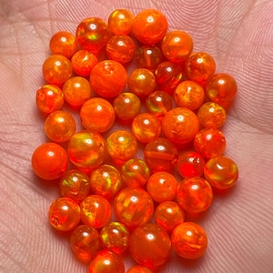 May include: A collection of vibrant orange opal beads in various sizes. The round gemstones display a fiery, iridescent glow, with flashes of yellow and red. The opals are held in a hand, showcasing their polished, spherical shapes.