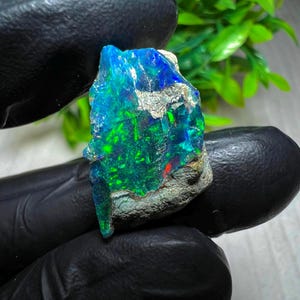 Puede incluir: Una vibrante piedra preciosa de ópalo en bruto con una llamativa exhibición de color. La piedra presenta una mezcla de tonos azules, verdes y rojos, con una superficie rugosa y texturizada. El ópalo se sostiene sobre un fondo oscuro.