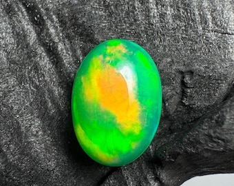 Pode incluir: Gema oval com uma paleta de cores vibrantes. A pedra apresenta uma base verde brilhante com manchas amarelas e laranja. A gema é colocada sobre um fundo escuro e texturizado, realçando o seu brilho.