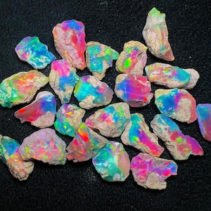 Può includere: Una collezione di gemme di opale grezze, che mostrano vivaci colori iridescenti. Gli opali presentano uno spettro di tonalità, tra cui blu, verdi, rosa e viola, su una matrice rocciosa neutra. Le gemme sono di varie dimensioni e forme.