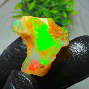 Könnte beinhalten: Ein roher Opal-Edelstein mit leuchtenden Farben. Der Opal zeigt einen hellgrünen Kern, umgeben von orangefarbenen und gelben Farbtönen. Der Stein hat eine raue, natürliche Textur und ist vor einem dunklen Hintergrund platziert.