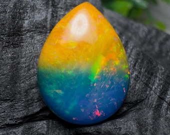 Pode incluir: Uma pedra preciosa de opala em forma de gota com um gradiente de cores vibrantes. A metade superior é amarelo brilhante, passando para um azul profundo na parte inferior. A pedra exibe flashes de verde e vermelho, em um fundo escuro.