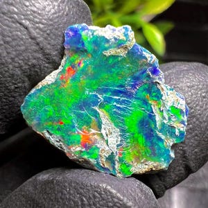 May include: A vibrant, raw opal stone with a striking display of color. The gemstone features a mix of green, blue, and red hues, with a rough, textured surface. The opal's colors shift and shimmer, creating a unique and eye-catching appearance.