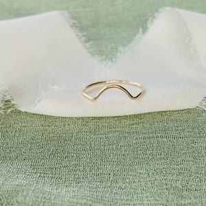 May include: A simple gold ring with a curved, wave-like design. The ring is on a white fabric background.