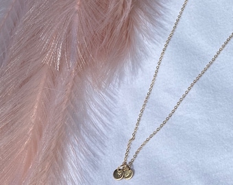 May include: A gold chain necklace with two small gold discs with letters on them. The necklace is on a white surface with a pink feather in the background.