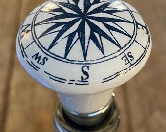 May include: White ceramic bottle stopper with a blue and white compass design. The compass features a star in the centre and the directions N, S, E, and W.