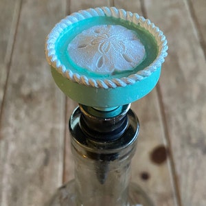 May include: A teal blue bottle stopper with a white rope design and a white sand dollar design in the centre.