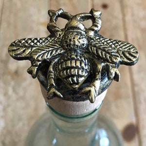 May include: A gold-toned metal bottle stopper shaped like a bee with detailed wings and a textured body.
