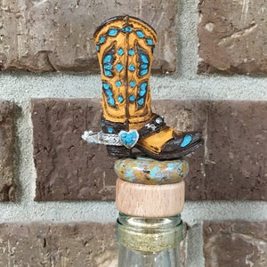 May include: A brown and yellow cowboy boot bottle stopper with turquoise accents and a silver spur. The boot is sitting on a wooden cork and a turquoise and brown patterned base.