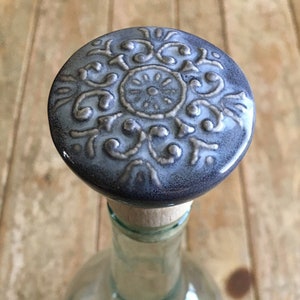 Puede incluir: Un tapón de botella decorativo con una parte superior de cerámica. La parte superior es de color azul grisáceo oscuro con un diseño floral en relieve detallado. El tapón es de corcho y está unido a una botella de vidrio transparente.