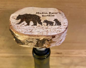 Może przedstawiać: Drewniany korek do butelki z motywem rodziny niedźwiedzi. Tekst "Mama Bear's Bottle" znajduje się nad ilustracją rodziny niedźwiedzi. Korek wykonany jest z naturalnego plastra drewna z krawędziami kory.