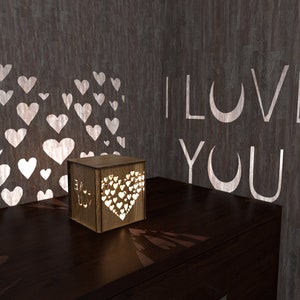 May include: A wooden box with a heart-shaped cutout that projects a pattern of hearts onto the wall. The box is lit from within and says "I Love You" on the side. The wall behind the box is a dark gray with a pattern of white hearts.